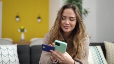 Akıllı telefon ve kredi kartı kullanan genç, güzel İspanyol kadın evdeki koltukta oturuyor.