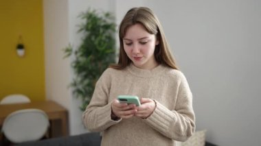 Young blonde woman using smartphone doing ok gesture at home