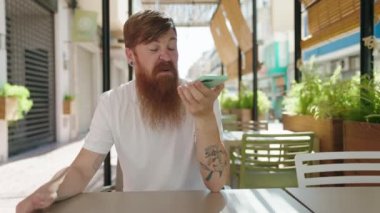 Young redhead man talking on the smartphone sitting on table at coffee shop terrace