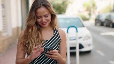 Genç, güzel, İspanyol bir kadın sokakta akıllı telefon kullanarak gülümsüyor.