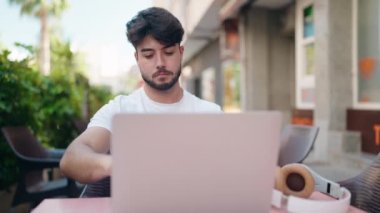 Young hispanic man using laptop sitting on table at coffee shop terrace