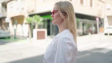 Genç sarışın kadın gülümsüyor. Kollarını kavuşturup caddede duruyor.