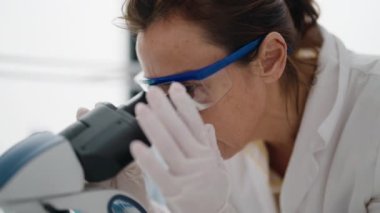 Laboratuvarda mikroskop kullanan orta yaşlı kadın bilim adamı üniforması giyiyor.