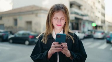 Beyaz bir kadın sokakta akıllı telefon kullanarak gülümsüyor.