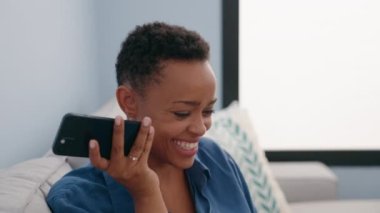 Afro-Amerikalı kadın akıllı telefondan evde kanepede oturmuş sesli mesaj dinliyor.