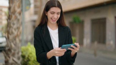 Genç İspanyol kız gülümsüyor. Sokakta akıllı telefondan videolar izliyor.