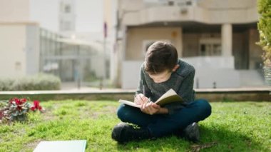 Blond child writing on book sitting on grass at park