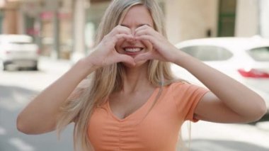 Young blonde woman smiling confident doing heart gesture with hands at street