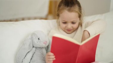Adorable blonde girl reading book sitting on bed at bedroom