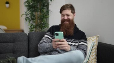 Young redhead man using smartphone sitting on sofa at home
