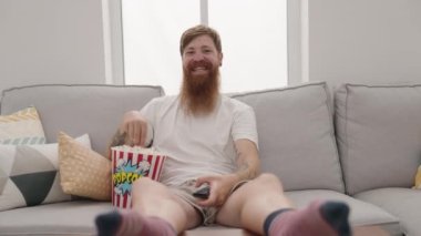 Young redhead man watching movie sitting on sofa at home