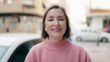 Orta yaşlı kadın gülümsüyor. Kollarını kavuşturup caddede duruyor.