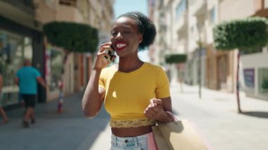 Afro-Amerikalı kadın akıllı telefonuyla konuşuyor. Elinde alışveriş torbalarıyla sokakta yürüyor.