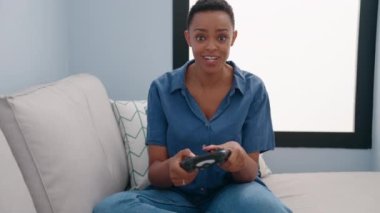 Evdeki kanepede oturan Afrikalı Amerikalı kadın video oyunu oynuyor.