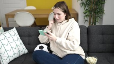 Young blonde woman watching soccer match on smartphone with nervous expression at home