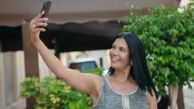 Orta yaşlı İspanyol kadın gülümsüyor. Sokakta akıllı telefonun yanında özçekim yapıyor.