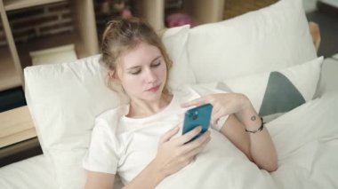 Yatak odasında mutsuz bir ifadeyle akıllı telefon kullanan genç sarışın kadın.
