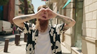 Genç kızıl saçlı adam gülümsüyor. Kendine güveni tam. Sokakta elleriyle kalp hareketi yapıyor.