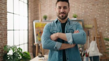 Genç İspanyol adam gülümsüyor. Kollarını kavuşturup sanat stüdyosunda duruyor.