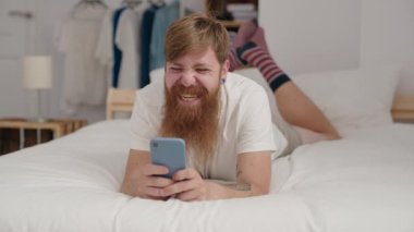 Genç kızıl saçlı adam yatakta akıllı telefon kullanıyor.