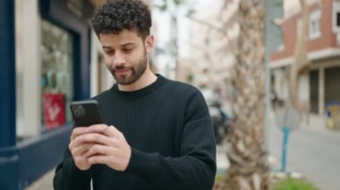 Genç Arap adam sokakta akıllı telefon kullanarak kendinden emin gülümsüyor.