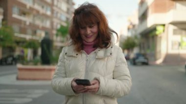 Middle age redhead woman smiling confident using smartphone at street