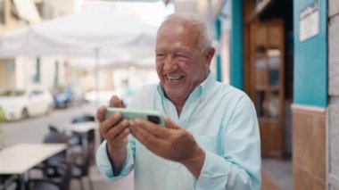 Senior grey-haired man smiling confident watching video on smartphone at street