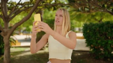 Genç sarışın kadın, kendinden emin gülümsüyor. Parktaki akıllı telefonun yanında fotoğraf çekiyor.