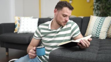Genç beyaz adam kanepede oturmuş kitap okuyor. Evde kahve içiyor.