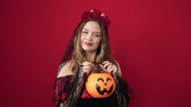 Young beautiful hispanic woman wearing katrina costume holding halloween pumpkin basket over isolated red background