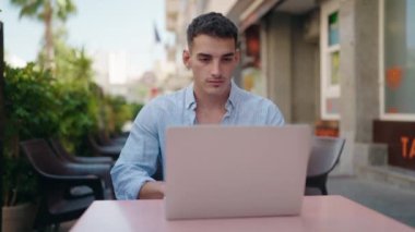 Young hispanic man using laptop sitting on table at coffee shop terrace