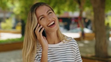 Genç sarışın kadın gülümsüyor. Parktaki akıllı telefondan konuşuyor.