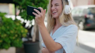 Genç sarışın kadın gülümsüyor. Sokakta akıllı telefondan videolar izliyor.