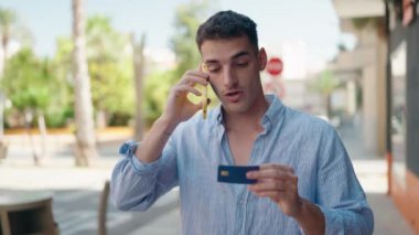 Young hispanic man talking on the smartphone and using credit card at street