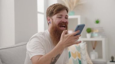 Young redhead man talking on the smartphone sitting on sofa at home
