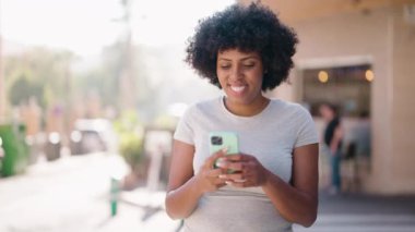 Genç Afrikalı Amerikalı kadın gülümsüyor. Kendine güveni tam. Sokakta akıllı telefondan selfie çekiyor.
