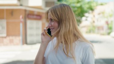 Genç sarışın kız gülümsüyor. Kendine güvenen. Sokakta akıllı telefondan konuşuyor.