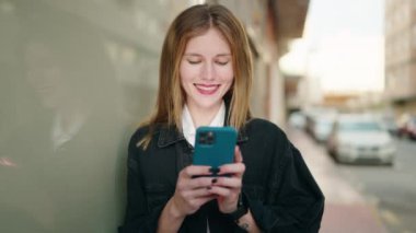 Beyaz bir kadın sokakta akıllı telefon kullanarak gülümsüyor.
