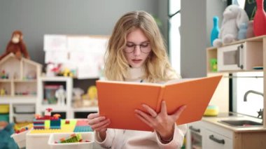 Young blonde woman preschool teacher reading book with relaxed expression at kindergarten