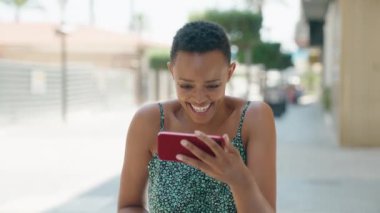 Afro-Amerikalı kadın gülümsüyor. Sokakta akıllı telefondan videolar izliyor.