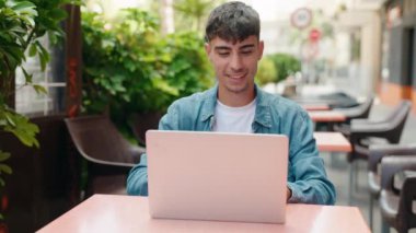Young hispanic man using laptop sitting on table at coffee shop terrace