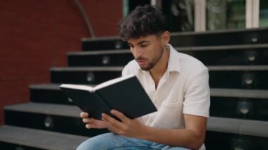 Young arab man reading book sitting on stairs at street
