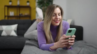 Evdeki kanepede uzanmış akıllı telefon kullanan genç sarışın kadın.
