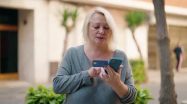 Middle age blonde woman using smartphone and credit card at street