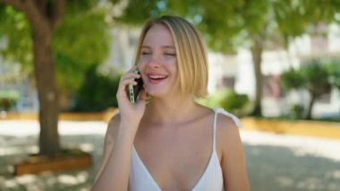 Genç sarışın kadın gülümsüyor. Parktaki akıllı telefondan konuşuyor.