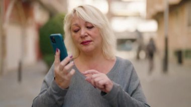 Orta yaşlı sarışın kadın sokakta akıllı telefondan konuşmaktan endişeleniyor.