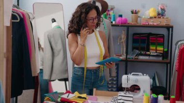 Middle age woman tailor talking on smartphone using touchpad at clothing factory