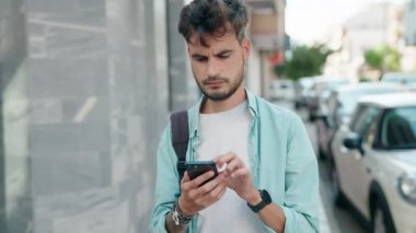 Young hispanic man student using smartphone with relaxed expression at street