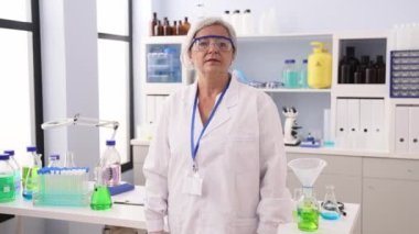 Orta yaşlı, gri saçlı, bilim adamı üniforması giyen bir kadın laboratuvarda kollarını kavuşturup el kol hareketi yapıyor.