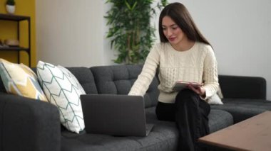 Young beautiful hispanic woman writing on notebook using laptop at home